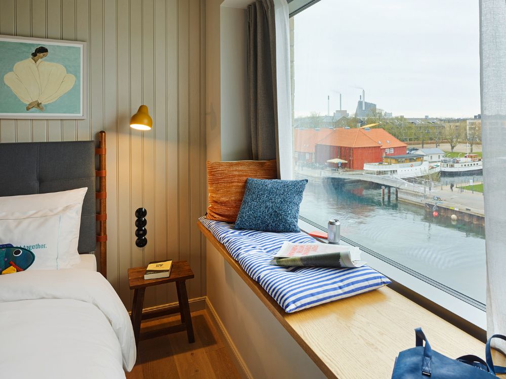 Cosy guest room bed next to a wide wooden window seat decorated with a blue-and-white striped cushion and pillows overlooking a city harbor.