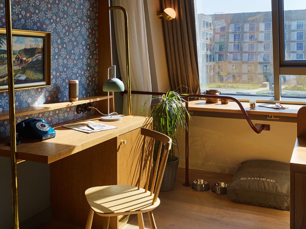 Chic guest room workspace featuring a wooden writing desk with a retro blue phone, a green lamp, and a floral wallpapered back panel.