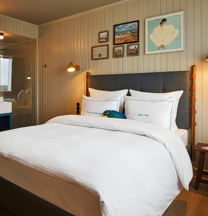 Serene hotel room featuring off-white vertical wood-paneled walls, framed artwork, a bed with a grey headboard, and a glimpse into a glass-enclosed shower.