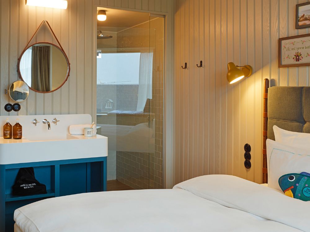 Modern guest room washbasin station featuring a white sink on a blue cabinet, a leather-strap round mirror, and a view into a glass-walled shower.