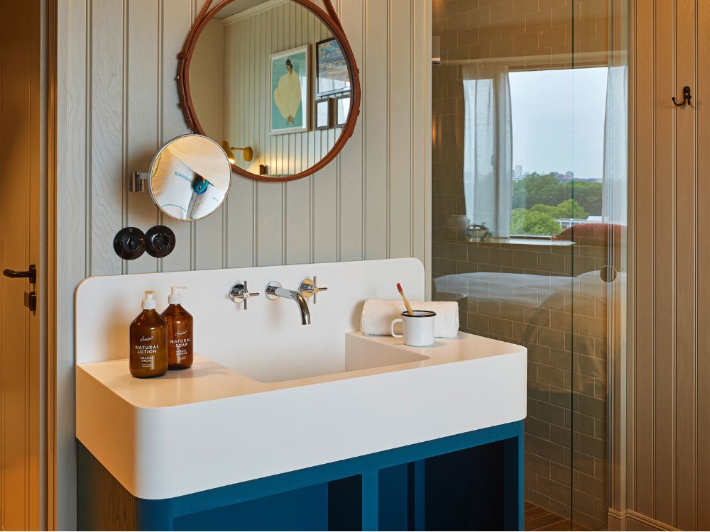 Modern guest room washbasin station featuring a white sink on a blue cabinet, a leather-strap round mirror, and a view into a glass-walled shower.