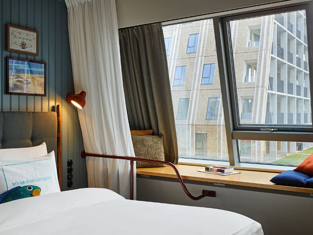 Close-up of a boutique hotel bed next to a large tilted window with a wooden sill used as a bench seat, showing a modern building outside.