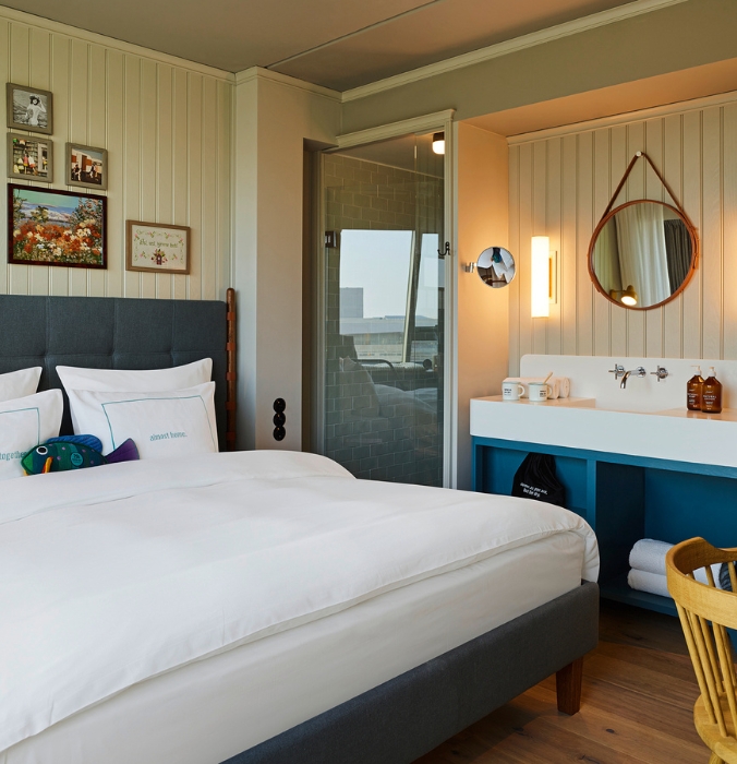 Comfortable hotel bed looking through to an open-concept washroom station featuring a white basin on a vibrant blue vanity cabinet and round leather-strap mirror.