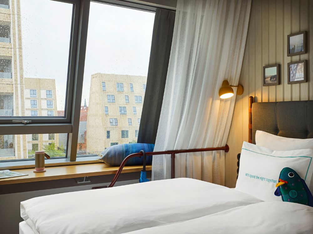 Boutique hotel bed with white linens next to a wide, slanted window with a wooden sill bench looking onto modern city buildings.