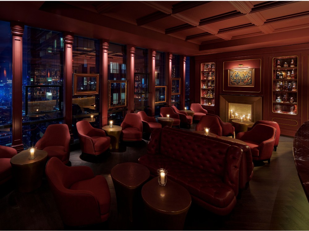 Cozy, dimly lit lounge with red armchairs, tufted sofa, candlelit tables, large windows showing cityscape, and a fireplace with shelves of bottles.