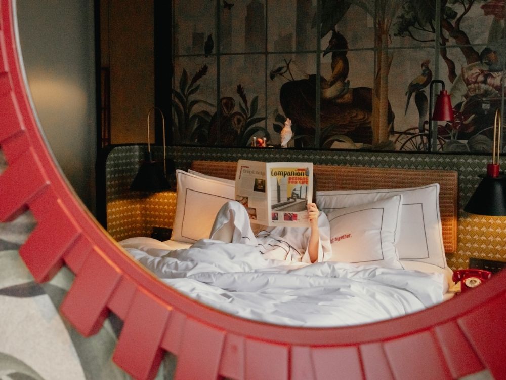 Person lying in bed reading a newspaper, reflected in a large circular red mirror; hotel room decor with artistic wall mural visible.