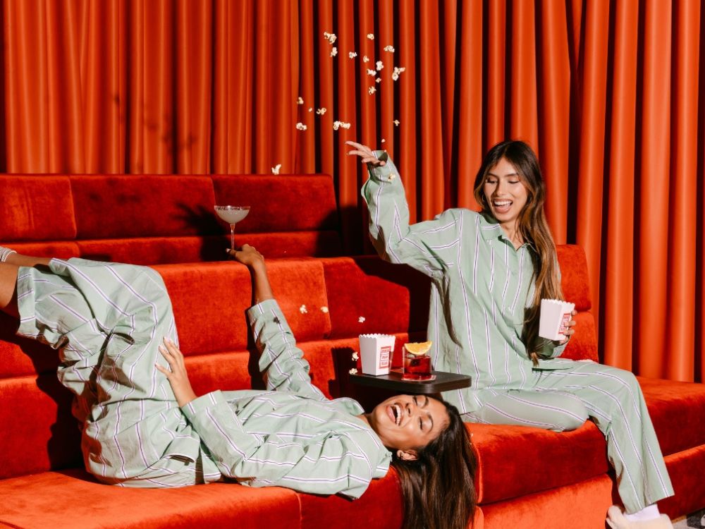 Two women in pajamas sit on a couch, enjoying popcorn and watching a movie together.