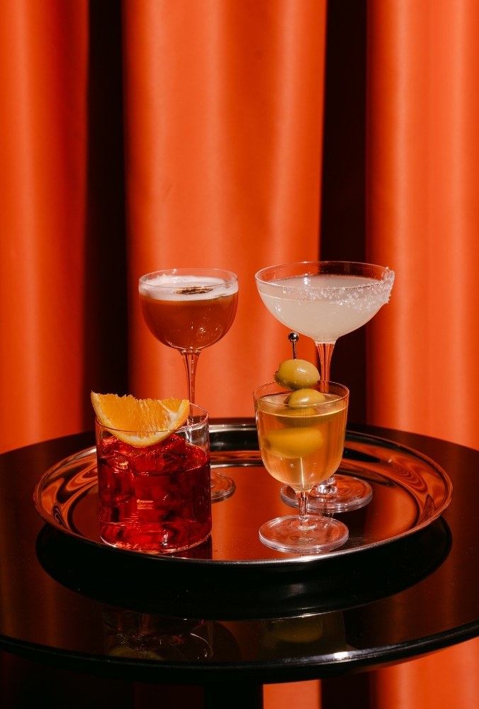 Four assorted cocktails on a silver tray against an orange curtain backdrop on a glossy black table.