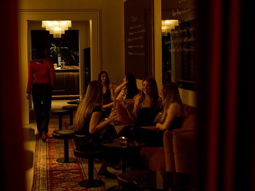 A group of women laughing and sharing drinks on a velvet sofa in a dimly lit, sophisticated hotel lounge with an oriental rug.