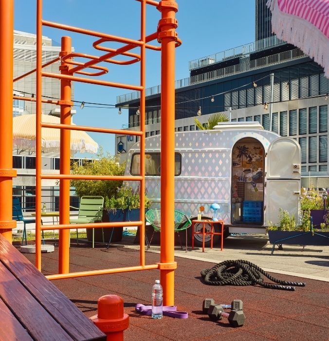 Outdoor rooftop workout area with orange calisthenics bars, dumbbells, battle ropes, and a pastel retro caravan.