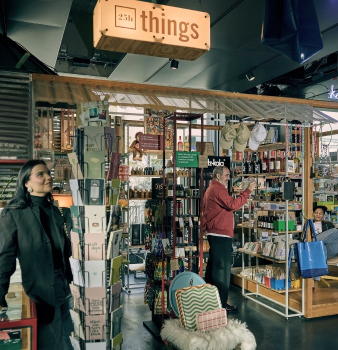 The colorful "25h things" concept store inside a 25hours Hotel lobby, with guests browsing a curated selection of postcards, books, design gifts, and travel accessories.