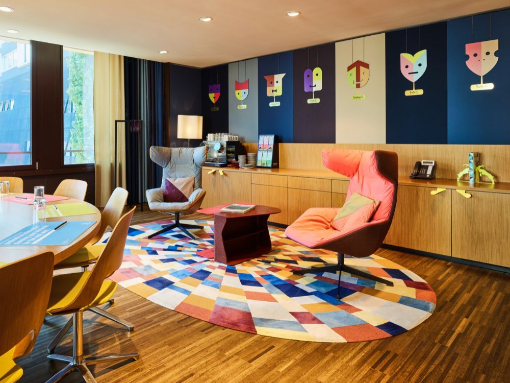 Colorful mid-century modern lounge with chairs, geometric rug, wood cabinetry, cocktail art, and warm sunlight creating a playful atmosphere.