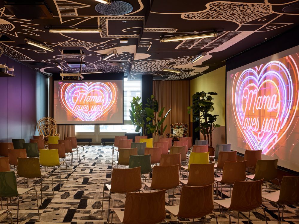 Modern lounge with patterned ceiling, colorful chairs, plants, and screens displaying glowing pink hearts and “Mama loves you,” creating a warm mood.