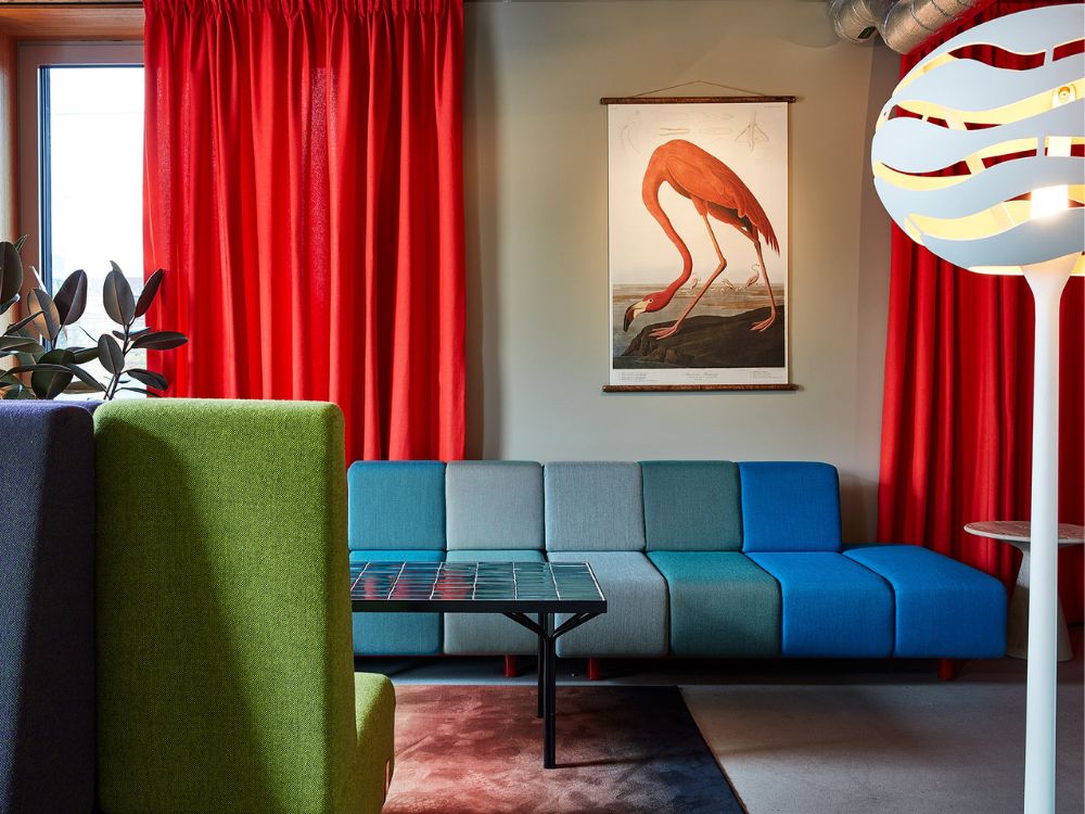 Retro-modern living room with vivid red curtains, blue sectional sofa, glass coffee table, flamingo artwork, and sculptural white lamp.