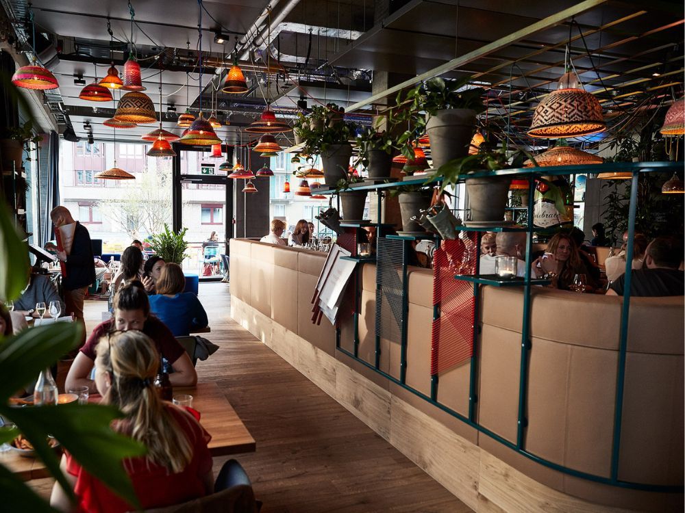 Lively, plant-filled restaurant interior with colorful pendant lights, diners at tables and booths, and sunlight streaming through large windows.