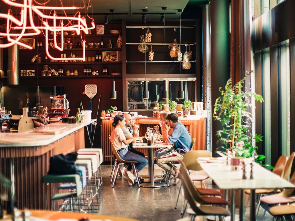 Stylish modern restaurant with a couple chatting over drinks, glowing red neon lights, warm wood accents, hanging glassware, and lush plants.