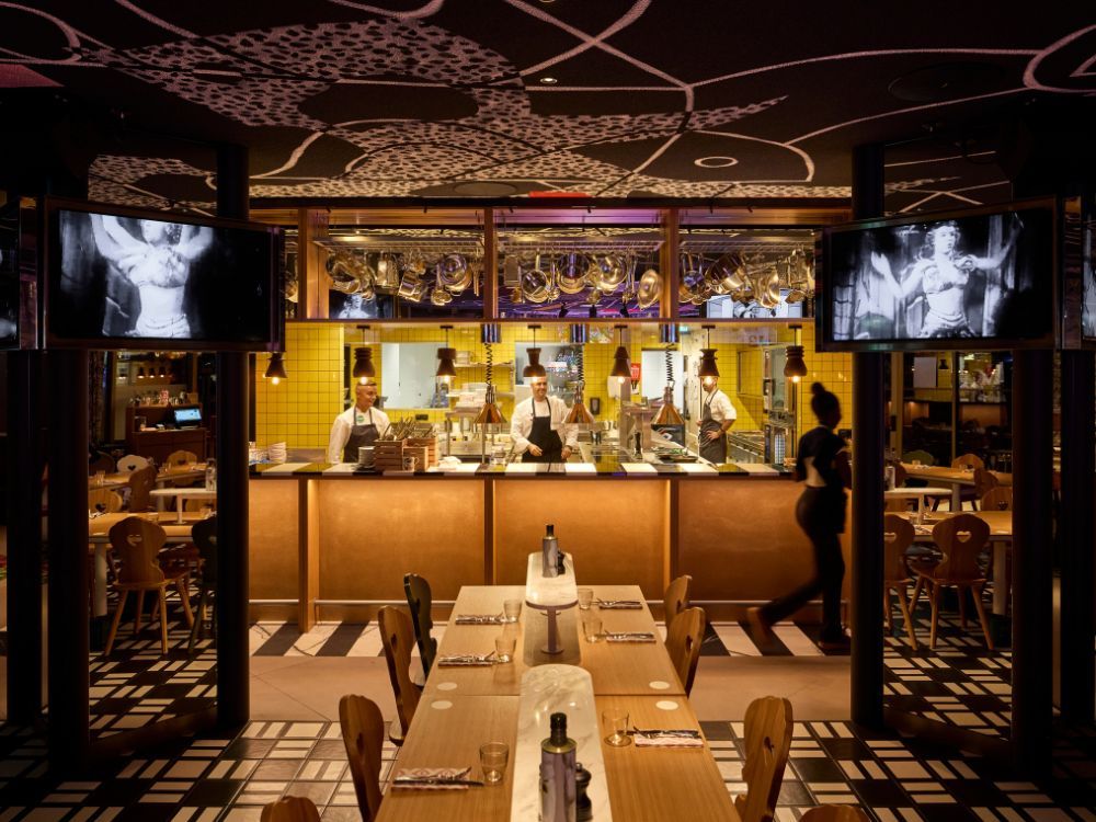 Upscale restaurant interior with chefs behind a glowing bar, set tables in front, ornate ceiling, and black-and-white screens; warm, lively mood.