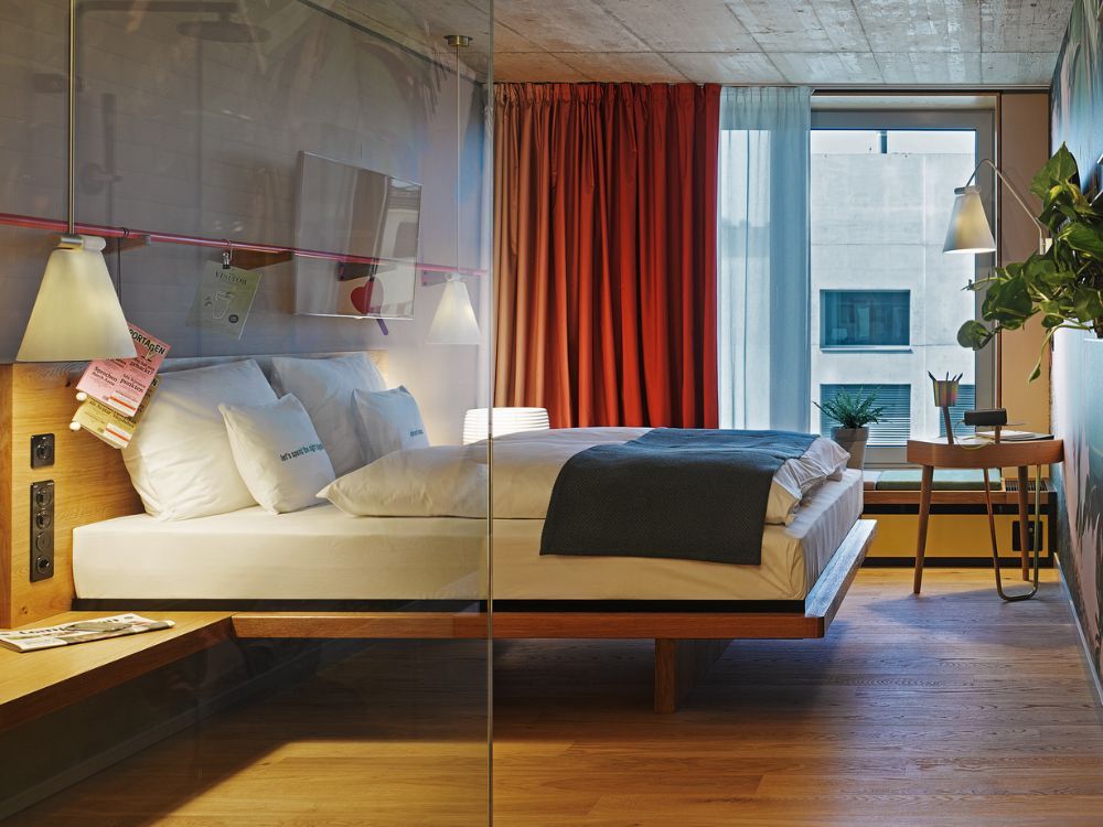 A modern hotel room featuring a wooden platform bed with white linens, red curtains, and a small desk by a window with plants.