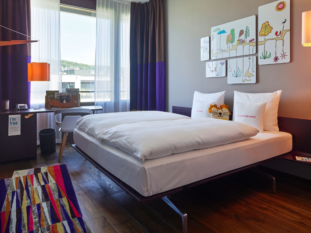 A modern hotel room features a bed with white linens, a colorful geometric rug, a desk, and whimsical wall art depicting trees and animals on a wall.