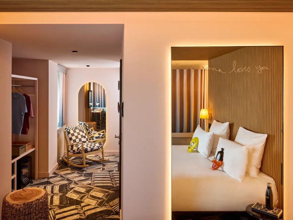 A hotel room features a bed with white linens, cartoon character pillows, and a wooden headboard, plus a seating area with checkered rocking chairs.