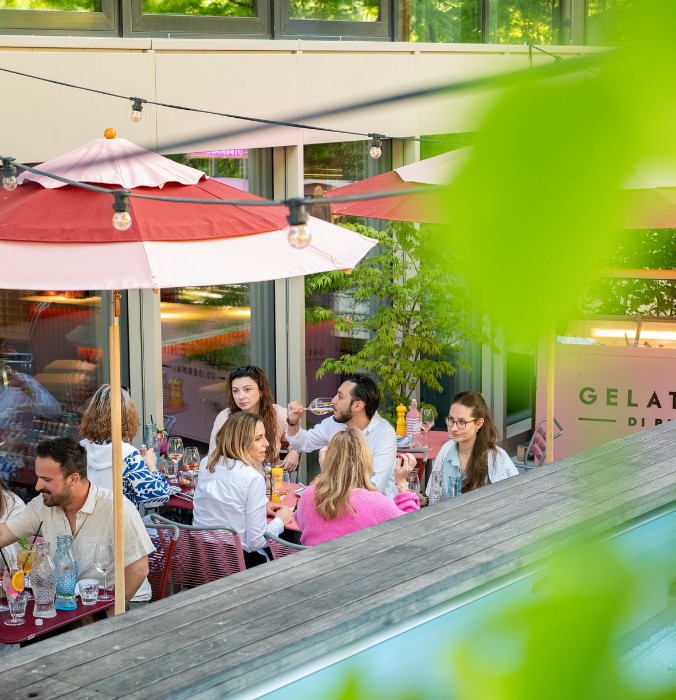 Group of people sitting at outdoor tables under umbrellas at a café with a gelato sign in the background