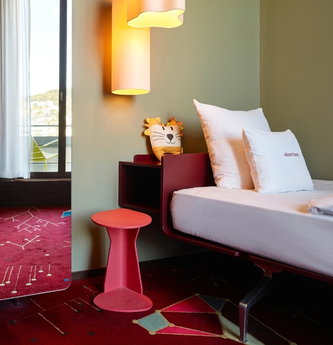 Playful hotel room featuring a modern single bed with a built-in dark red nightstand, a lion plush pillow, a pink wooden stool, and a vibrant patterned red carpet.