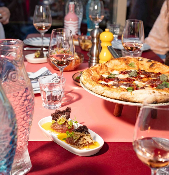 An elegant dinner table at Ribelli Italian restaurant featuring fresh stone-baked Neapolitan pizza, gourmet tuna tartare, and glasses of chilled rosé wine.