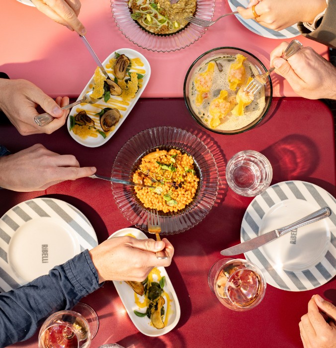 Overhead view of friends sharing a variety of colorful Italian dishes including fresh pasta, antipasti, and wine on a red dining table.