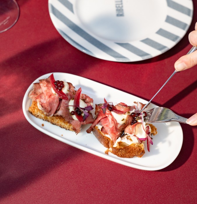 Two crispy beef bruschetta slices topped with cream sauce and savory balsamic pearls, being served on a red table.