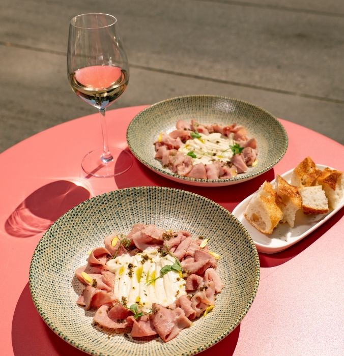 Classic Italian vitello tonnato served in ceramic bowls with a side of crusty bread and a glass of white wine on a pink outdoor table.