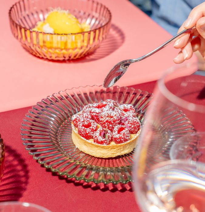 Close-up of a fresh raspberry tart dusted with powdered sugar, served in a clear glass dish next to a bowl of yellow mango sorbet.
