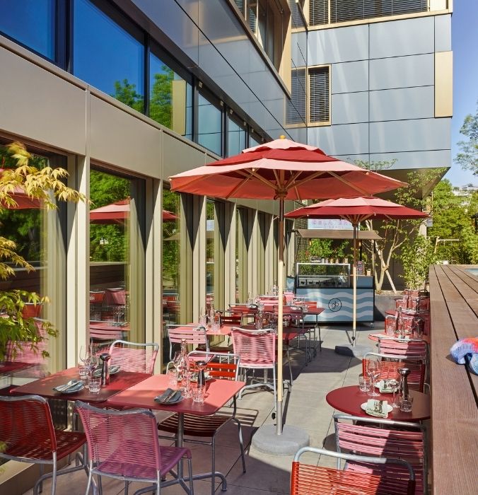 Sunny outdoor dining terrace of a boutique hotel restaurant featuring chic red tables, pink sun umbrellas, and a vintage gelateria cart.