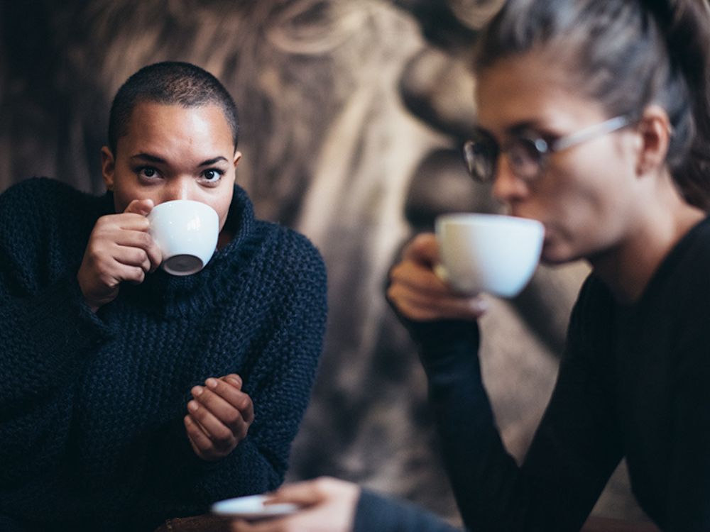 Two women wearing dark sweaters sit together in a dimly lit space, both holding white ceramic cups and drinking coffee while looking ahead intently.