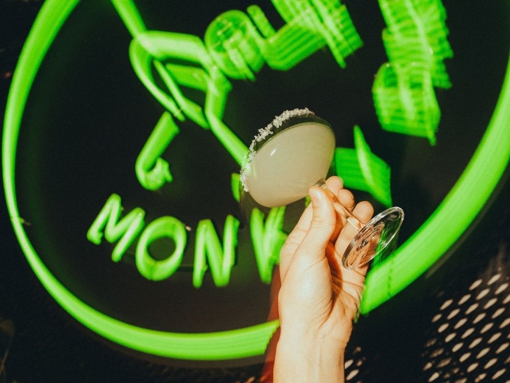 A hand holds a cocktail against a bright green neon sign.