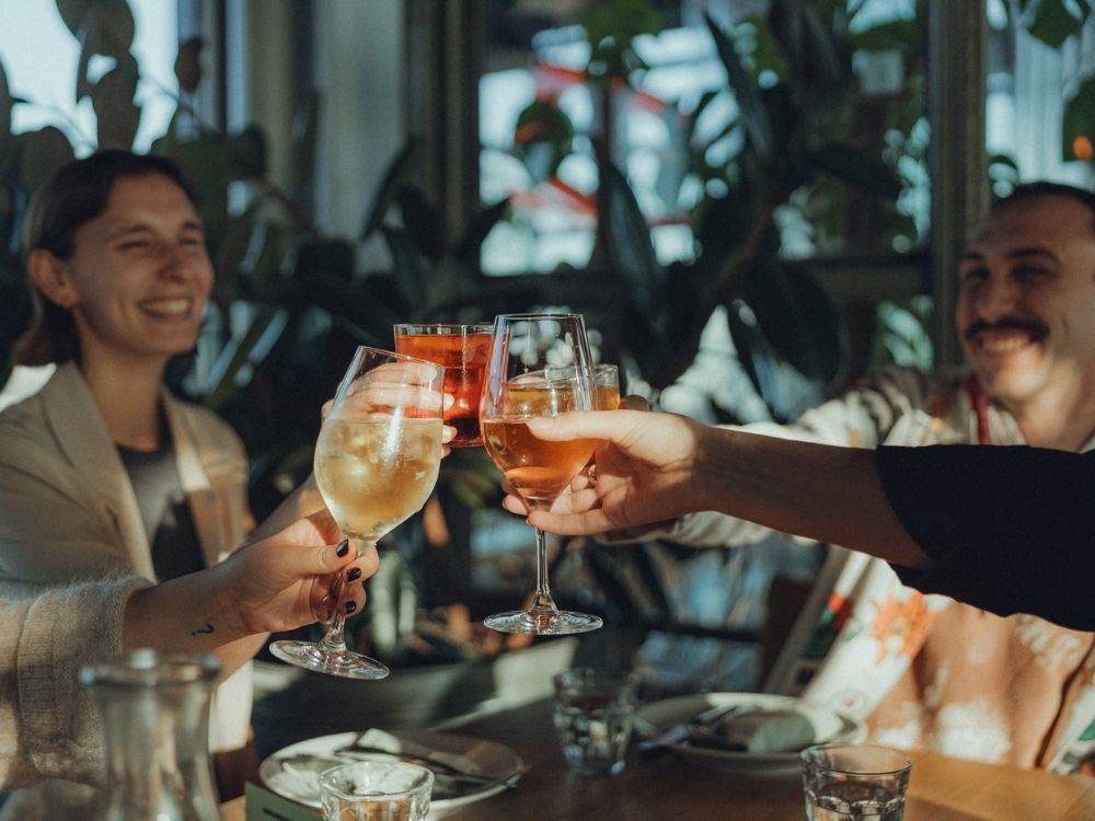 Four people clink glasses at a restaurant table, smiling and enjoying drinks. Plates, glasses, and green plants are visible in the background.