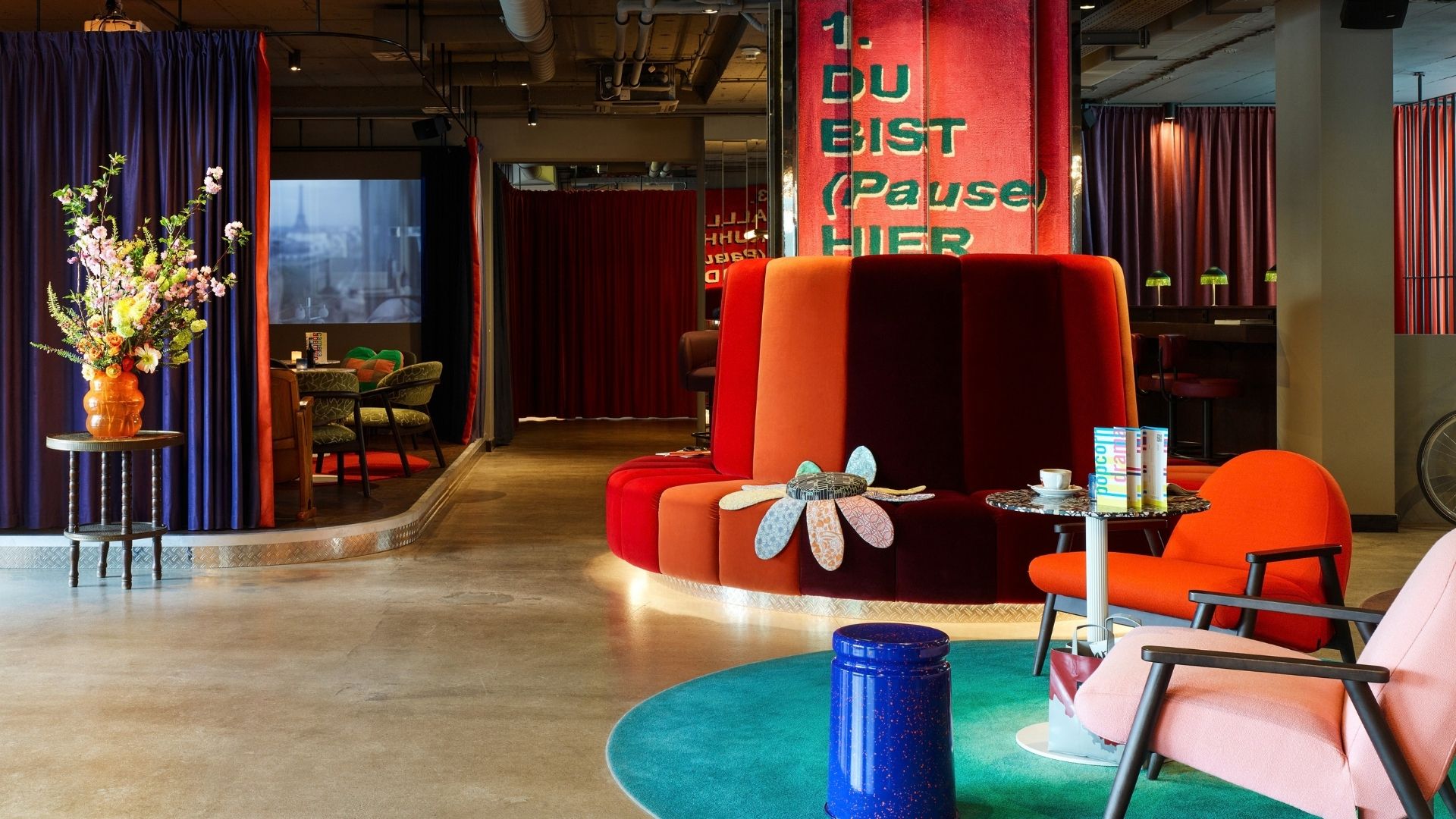 Vibrant modern lounge with a towering red sofa, orange and pink chairs, blue tables, flowers, and dramatic curtains, creating a playful, inviting mood.