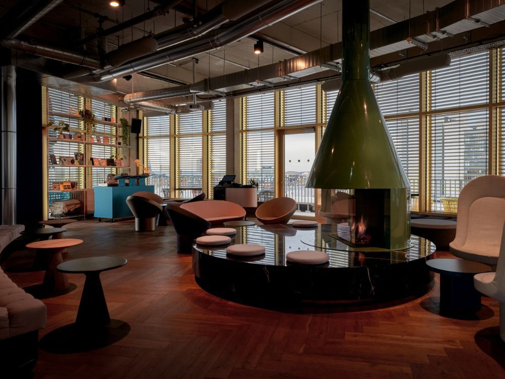 Modern hotel rooftop lounge with a prominent cone-shaped green fireplace, mid-century modern chairs, and parquet wooden flooring under industrial-style ceilings.