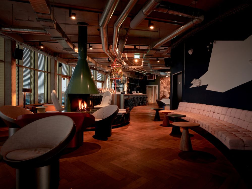 Retro-futuristic hotel lounge featuring white pod chairs, a green central fireplace, and large windows with blinds.