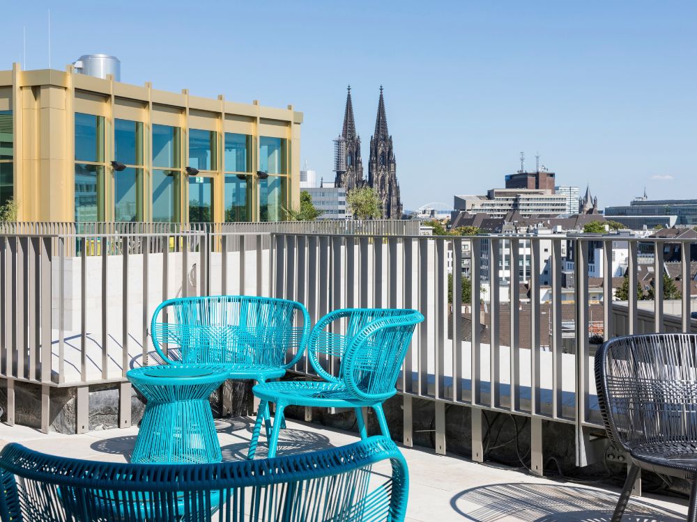 Rooftop terrace with bright turquoise woven chairs and table, featuring a clear view of the historic Cologne Cathedral in Germany.