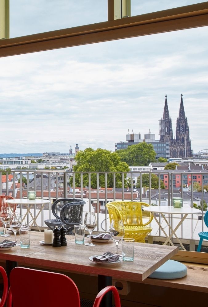 Rooftop restaurant dining table looking out to an outdoor balcony with colorful yellow and blue wicker chairs and a view of the Cologne Cathedral.