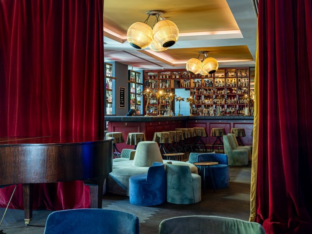 Elegant hotel bar and lounge with red velvet curtains, a grand piano, colorful velvet tub chairs, and a fully stocked back bar.