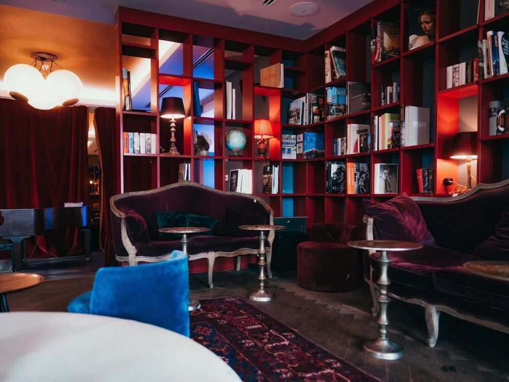 Cozy hotel library lounge with red floor-to-ceiling bookshelves, antique purple velvet sofas, brass side tables, and a traditional patterned rug.