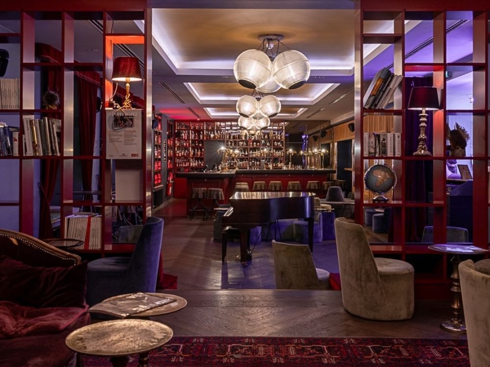 Elegant hotel lounge and jazz bar viewed through red wooden partitions, featuring a central black grand piano, low-profile modern armchairs, and a fully stocked back bar.