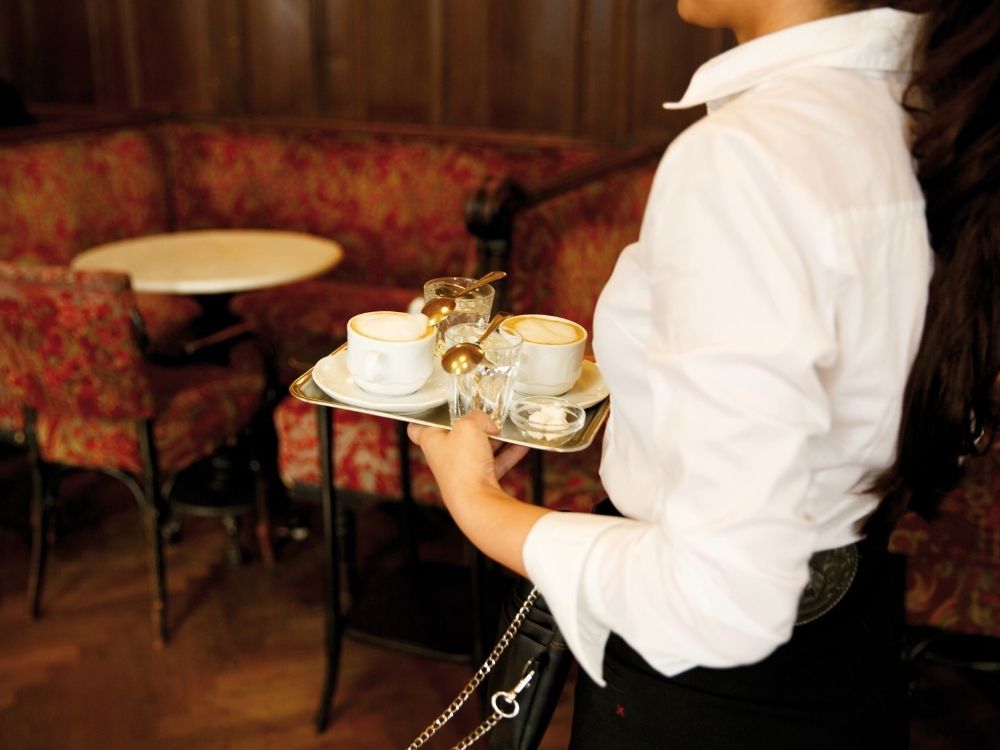 Waitress in a white shirt carrying a silver tray with traditional Viennese coffee and glasses of water in a classic Vienna coffee house.

