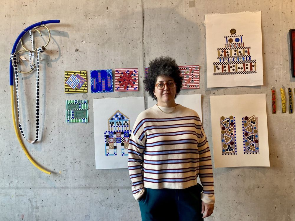 A woman stands in front of a colorful wall adorned with various pieces of art, showcasing her appreciation for creativity.