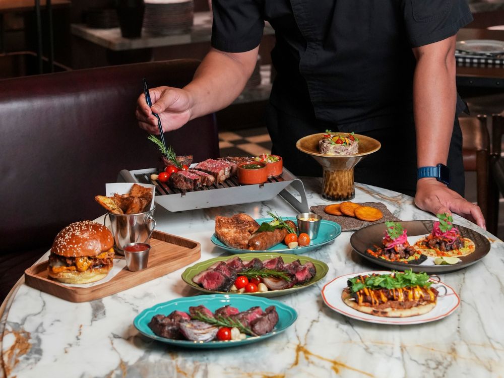 A chef presenting a gourmet feast with a variety of grilled meat dishes, including steaks, a burger, tacos, and sausages on a marble table.