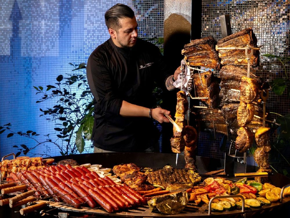 A chef preparing a large selection of meats on vertical skewers, sausages, and grilled vegetables at a professional barbecue station.