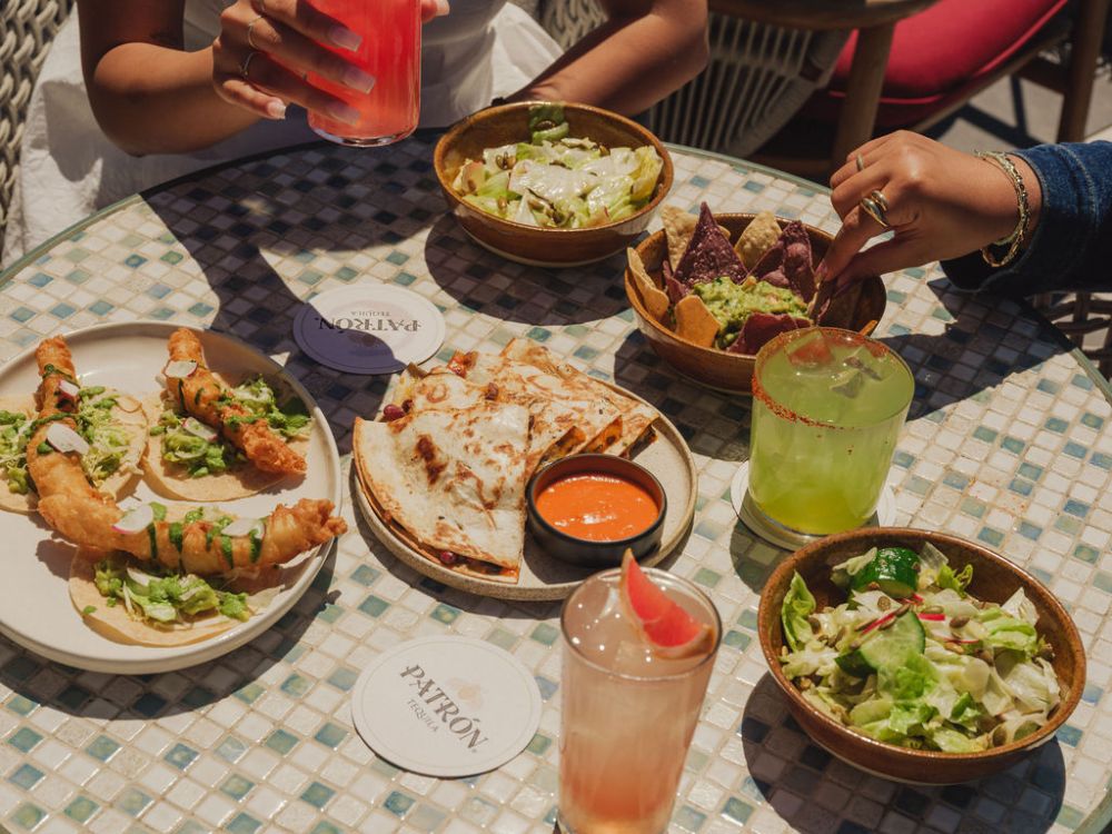 Overhead view of diners sharing tacos, salads, and dip with colorful cocktails on a sunlit mosaic table, creating a relaxed, lively dining mood.