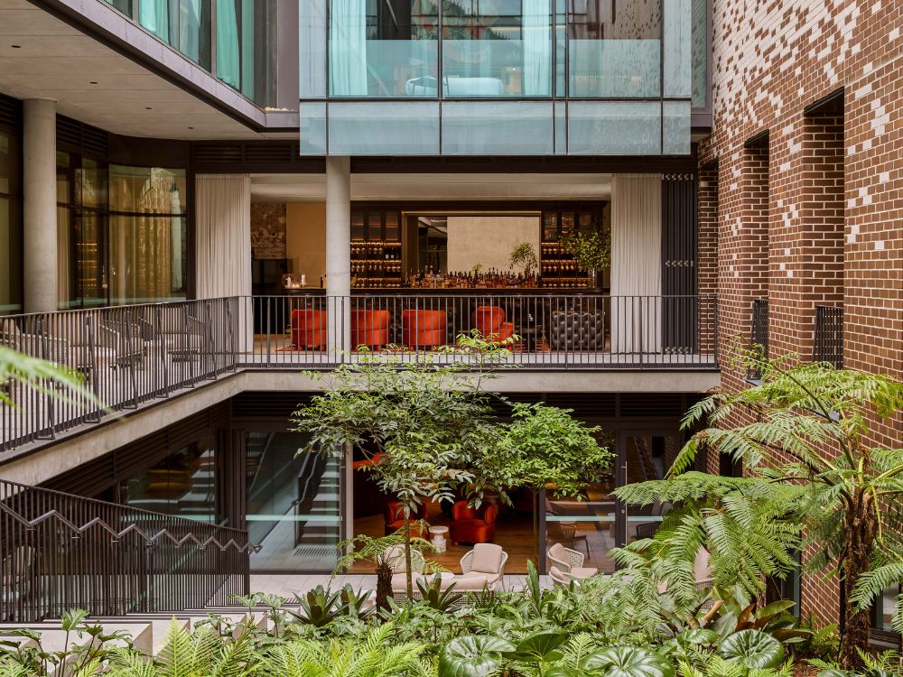 A modern brick-and-glass courtyard with layered balconies, red lounge chairs, and lush ferns and trees creates a calm, inviting urban retreat.