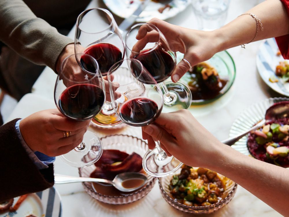 Four hands raise glasses of red wine for a toast over a dinner table filled with plated food, creating a warm, festive mood.